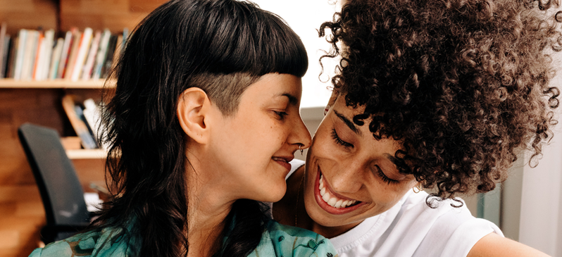 Two individuals posed side by side, each smiling. The person to the left has tan skin and a black mullet with blunt bangs and a septum piercing. They are facing the person to the right, who is leaning over their shoulder– they have dark skin and curly brown hair pulled into an updo.