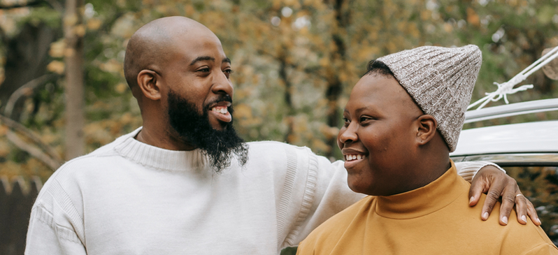 Two individuals posed next to each other, smiling. The person on the left has dark skin and a black beard, with their arm around the person to the right of them. The person to the right has dark skin and a gray beanie on, and is looking at the person to the left of them.