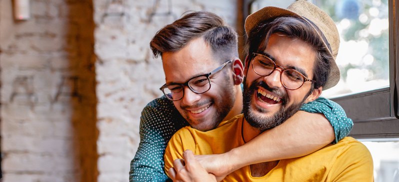 Two individuals hugging each other and smiling. The person further behind the other has glasses, combed over brunette hair, and has their arms around the person in front of them. The person in front of them has brown combed over hair, a beige hat, a beard, and glasses.